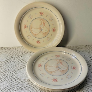 May include: Two cream-colored ceramic dinner plates featuring a central illustration of two geese wearing blue ribbons. The design includes a border of small pink and blue floral clusters, a blue dotted circle, and a thin blue rim line around the edge.