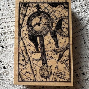 May include: A rectangular wooden stamp featuring a detailed black illustration of a clock pendulum and a celestial map. The text "HOROLOGIUM PENDULUM" is visible. The stamp has a vintage, antique aesthetic.