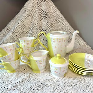 May include: A vintage tea set with a white and yellow color scheme. The set includes a teapot, creamer, sugar bowl, saucers, and teacups. The teacups, creamer, and teapot have a yellow stripe design. The set is decorated with small pink floral patterns.