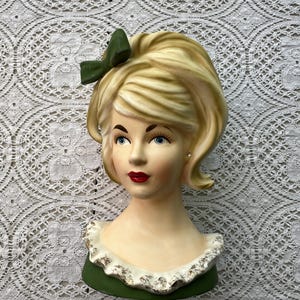 May include: A vintage ceramic wall vase in the shape of a woman's head. The blonde hair is styled in a bouffant with a green bow. The face has blue eyes and red lips. The green base has a white and gold trim.