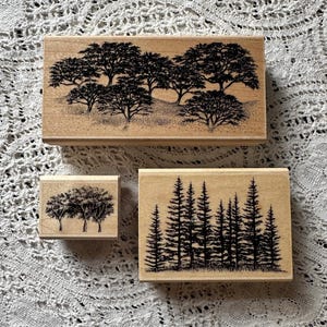 May include: Three wooden stamps with black tree designs. The largest stamp features a group of trees on a hill, the middle stamp has three trees, and the smallest stamp has a forest of pine trees.