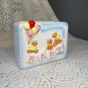 May include: A rectangular ceramic baby-themed planter. The planter is light blue with a white border and features embossed teddy bears, balloons, and the word "BABY" in pink letters. The balloons are red, yellow, and pink.