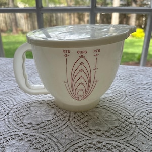 May include: Off-white vintage mixing bowl with a matching lid and handle. The bowl has red measurement markings for quarts, cups, and pints. The bowl is sitting on a white lace tablecloth.