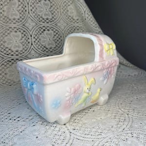 May include: A ceramic baby cradle planter in pastel pink, blue, and yellow. The planter features embossed floral and animal designs, including a bunny and duckling. A blue bow adorns the side, and a yellow bow accents the hood.