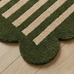 May include: A tufted rug featuring a scalloped edge design with alternating thick stripes in forest green and cream. The textile surface has a dense, textured pile construction that creates a soft appearance across the entire patterned area.