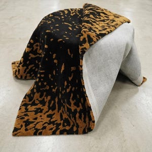 May include: A black and tan patterned rug is draped over an unseen object. The rug's design features a repeating pattern of tan shapes against a black background. The underside of the rug is a light gray color, revealing its construction.
