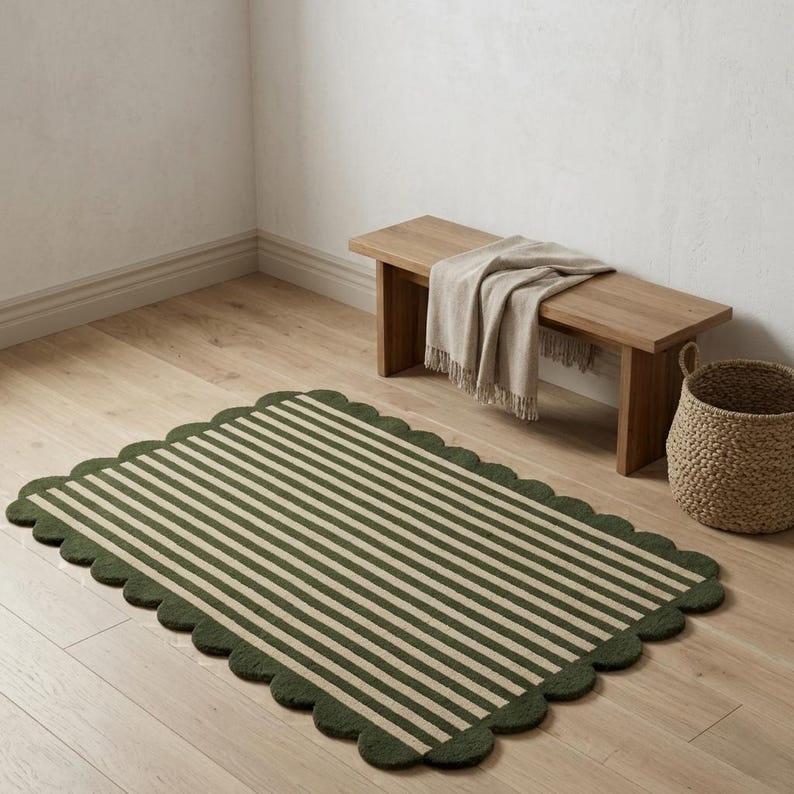 May include: A rectangular rug featuring a pattern of alternating dark green and cream-coloured stripes with a scalloped edge. The rug is positioned on a light-coloured wooden floor near a wooden bench draped with a beige throw and a woven basket.