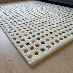 Könnte beinhalten: Cremefarbener Teppich mit einem Rastermuster aus kreisförmigen Aussparungen. Die Aussparungen sind mit verschiedenen Farben gefüllt, darunter Rot, Blau, Grün und Gelb. Der Teppich besteht aus einem weichen, strukturierten Material.