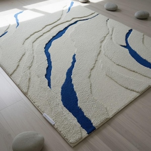 May include: A rectangular off-white rug with a textured surface and blue river-like patterns. The rug is placed on a light wood floor, with three gray stones nearby. The rug has a tag on the side.