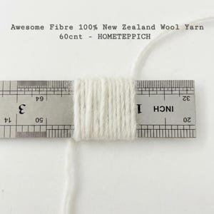 May include: Off-white wool yarn wrapped around a metal ruler, showing a thickness of approximately 1.3 cm. The text Awesome Fibre 100% New Zealand Wool Yarn 60cnt - HOMETEPPICH is printed above the yarn and ruler on a plain white background.