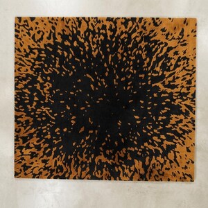 May include: Square rug with a black center and a golden-brown, speckled pattern radiating outwards. The rug's design resembles an abstract burst or explosion, creating a striking visual contrast. The rug is likely made of a soft material.