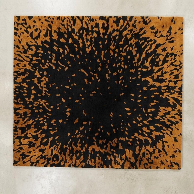May include: Square rug with a black center and a golden-brown, speckled pattern radiating outwards. The rug's design resembles an abstract burst or explosion, creating a striking visual contrast. The rug is likely made of a soft material.