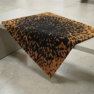 May include: A rectangular rug with a black center and a brown, speckled border. The rug is placed on a white table, with the corner of the rug extending over the edge. The rug's design has a textured appearance.