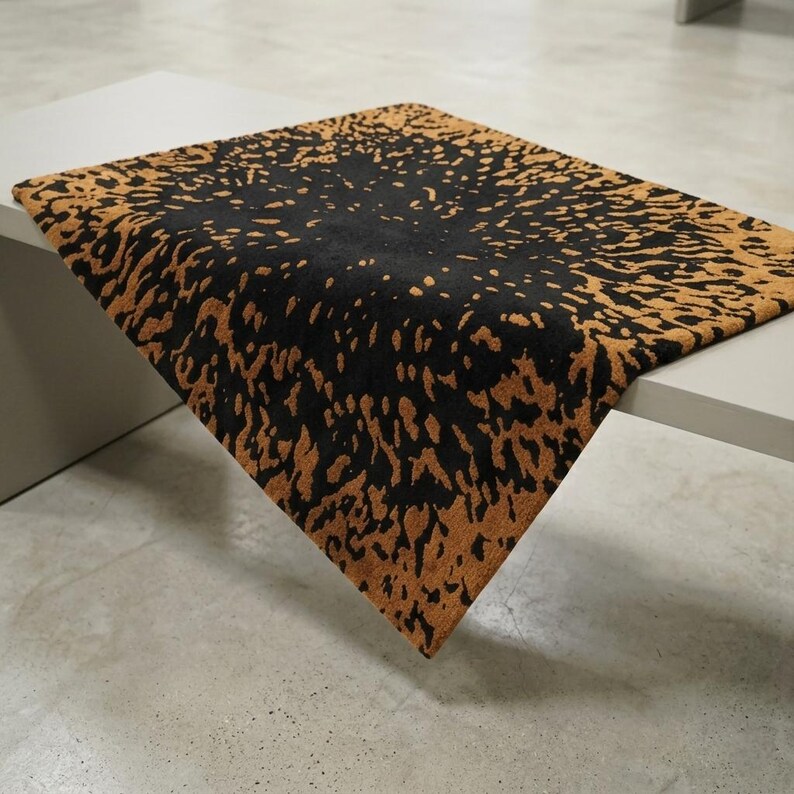 May include: A rectangular rug with a black center and a brown, speckled border. The rug is placed on a white table, with the corner of the rug extending over the edge. The rug's design has a textured appearance.