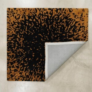 May include: A square rug with a black and gold abstract pattern. The rug's design features a dark black center with a gold-colored, irregular pattern that fades towards the edges. The corner is folded back, revealing a gray underside.