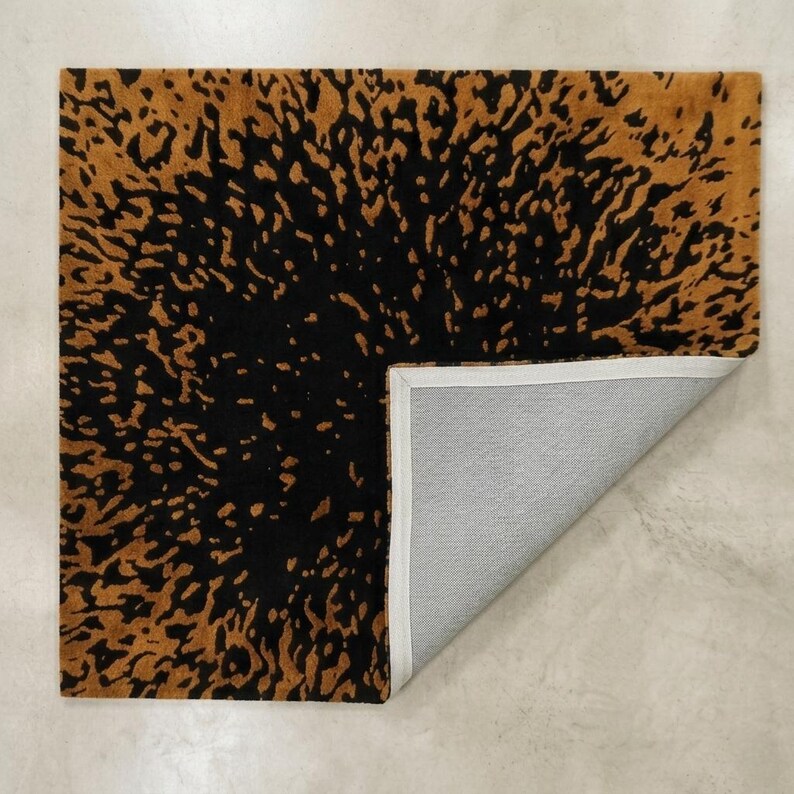 May include: A square rug with a black and gold abstract pattern. The rug's design features a dark black center with a gold-colored, irregular pattern that fades towards the edges. The corner is folded back, revealing a gray underside.
