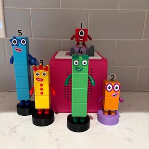 May include: A set of Numberblocks figures, numbered one to five. The figures are in various colours, including blue, yellow, green, orange, and red. They stand on black or purple bases, with a pink box in the background. Educational toys.