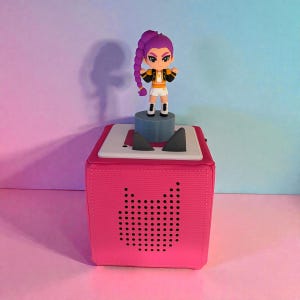 May include: A bright pink audio player with a cat-shaped speaker grille. A small figurine of a character with purple hair and a black and gold jacket stands on top of the player.