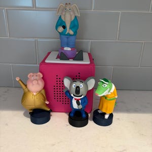 Puede incluir: Una caja de altavoz rosa con cuatro figuras de personajes de la película "Sing". Las figuras incluyen un elefante, un cerdo, un koala y un cocodrilo. Cada personaje sostiene un micrófono y está de pie sobre una base negra.