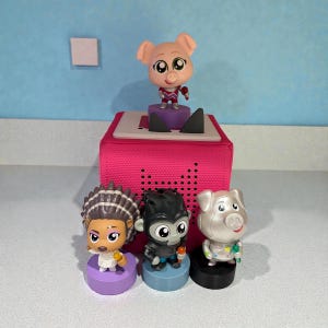 May include: A pink audio player with a pig figurine on top and three other character figurines in front. The figurines are a pig, a gorilla, and a woman. The figurines are on colored bases.
