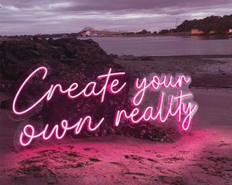 May include: A pink neon sign with the words "Create your own reality" in a cursive font. The sign is on a beach with a body of water and a bridge in the background. The sky is overcast with shades of purple and gray.