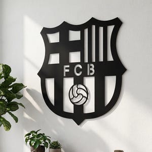May include: Black metal wall art featuring the FC Barcelona crest. The shield-shaped emblem includes the letters "FCB" and a soccer ball. The artwork is mounted on a white wall in a modern living space with a wooden cabinet and plants.