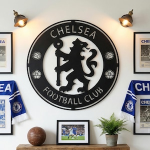 May include: A wall display featuring a large black Chelsea Football Club emblem, framed vintage posters, a scarf, and a vintage soccer ball. The emblem is centered, with the team name and a lion crest. Two wall-mounted lights illuminate the display.