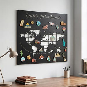 May include: Black world map with white grid lines and travel-themed magnets. The text "Amily's Global Travels" is written in silver script at the top. Magnets include landmarks, vehicles, and travel icons. The map is mounted on a wall above a wooden desk.