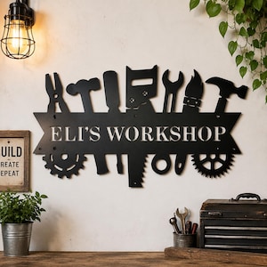 Peut inclure: Une enseigne murale en métal noir avec le texte ELI'S WORKSHOP sur une bannière centrale. Le design présente des silhouettes d'outils variés comme des pinces, un marteau, un tournevis, une scie, une clé, un pinceau et des engrenages sur les bords.