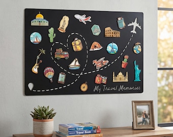 Puede incluir: Pizarra magnética negra con imanes temáticos de viajes, incluyendo monumentos y vehículos. La pizarra muestra el texto "My Travel Memories" y se exhibe en una pared sobre un mueble de madera con una planta en maceta y libros.