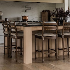 Op de afbeelding: Een set van vier donkerbruine houten barkrukken met geschulpte rugleuningen en beige gestreepte stoffen zitkussens vastgemaakt met dunne koorden, geplaatst rond een groot kookeiland met een zwart aanrechtblad en licht houten kasten.