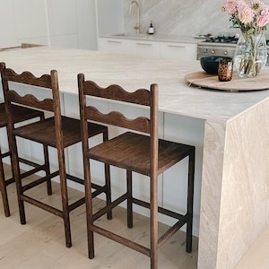 K&ouml;nnte beinhalten: Zwei dunkelbraune Holz-Barhocker mit vier geraden Beinen, einer quadratischen Sitzfl&auml;che und einer R&uuml;ckenlehne mit einer markanten, wellenf&ouml;rmigen oberen Leiste. Die Hocker sind mit horizontalen St&uuml;tzstreben zwischen den Beinen f&uuml;r Stabilit&auml;t konstruiert.