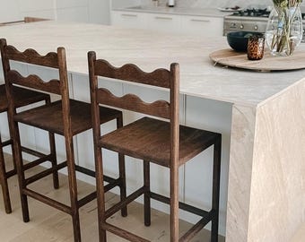 May include: Two dark brown wooden bar stools with four straight legs, a square seat, and a backrest featuring a distinct scalloped top rail. The stools are constructed with horizontal support bars between the legs for stability and durability.