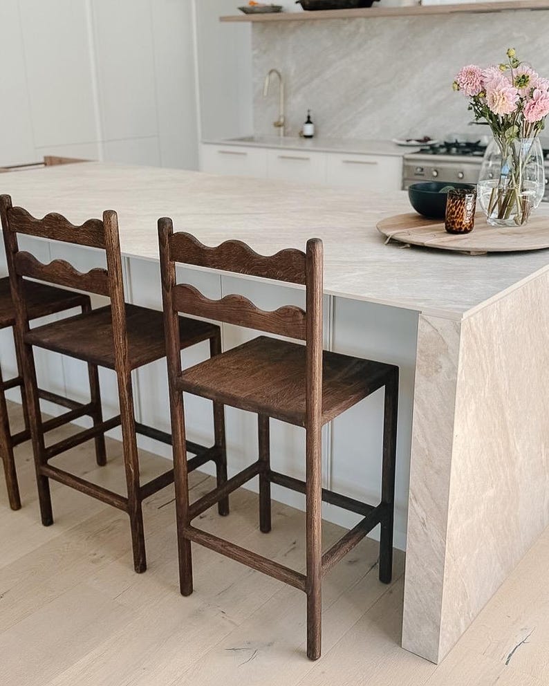 K&ouml;nnte beinhalten: Zwei dunkelbraune Holz-Barhocker mit vier geraden Beinen, einer quadratischen Sitzfl&auml;che und einer R&uuml;ckenlehne mit einer markanten, wellenf&ouml;rmigen oberen Leiste. Die Hocker sind mit horizontalen St&uuml;tzstreben zwischen den Beinen f&uuml;r Stabilit&auml;t konstruiert.