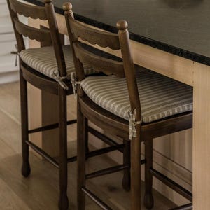 Könnte beinhalten: Zwei dunkelbraune Holz-Barhocker mit geschwungenen Rückenlehnen und beigeweiß gestreiften Sitzkissen. Jedes Kissen ist mit Stoffbändern an den Seiten am Holzrahmen befestigt. Die Hocker verfügen über vier Beine und Fußstützen.