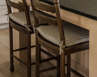 Könnte beinhalten: Zwei dunkelbraune Holz-Barhocker mit geschwungenen Rückenlehnen und vier Beinen. Jeder Hocker verfügt über ein beigeweiß gestreiftes Stoff-Sitzkissen, das mit Stoffbändern an den hinteren Ecken befestigt ist und eine gepolsterte Sitzfläche bietet.