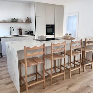 K&ouml;nnte beinhalten: Vier h&ouml;lzerne Barhocker mit geschwungenen R&uuml;ckenlehnen, die an einer K&uuml;cheninsel aus wei&szlig;em Marmor stehen. Die Hocker haben eine nat&uuml;rliche Holzmaserung, vier gerade Beine und eine horizontale St&uuml;tzstrebe f&uuml;r die F&uuml;&szlig;e.