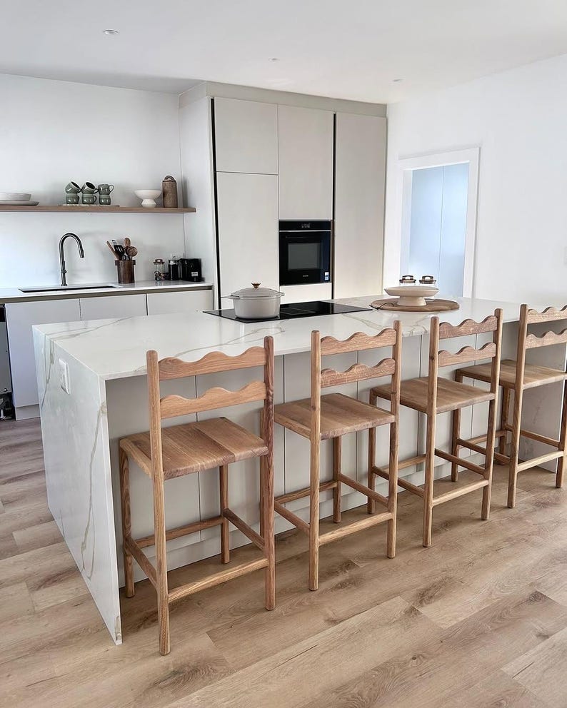 K&ouml;nnte beinhalten: Vier h&ouml;lzerne Barhocker mit geschwungenen R&uuml;ckenlehnen, die an einer K&uuml;cheninsel aus wei&szlig;em Marmor stehen. Die Hocker haben eine nat&uuml;rliche Holzmaserung, vier gerade Beine und eine horizontale St&uuml;tzstrebe f&uuml;r die F&uuml;&szlig;e.