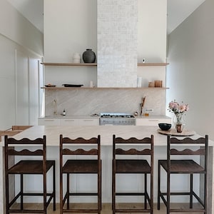 K&ouml;nnte beinhalten: Vier dunkelbraune Holzbarhocker mit geschwungenen R&uuml;ckenlehnen stehen in einer Reihe vor einer K&uuml;cheninsel mit einer hellen Steinarbeitsplatte. Die K&uuml;che verf&uuml;gt &uuml;ber wei&szlig;e Schr&auml;nke, einen gefliesten Fliesenspiegel und offene Holzregale.