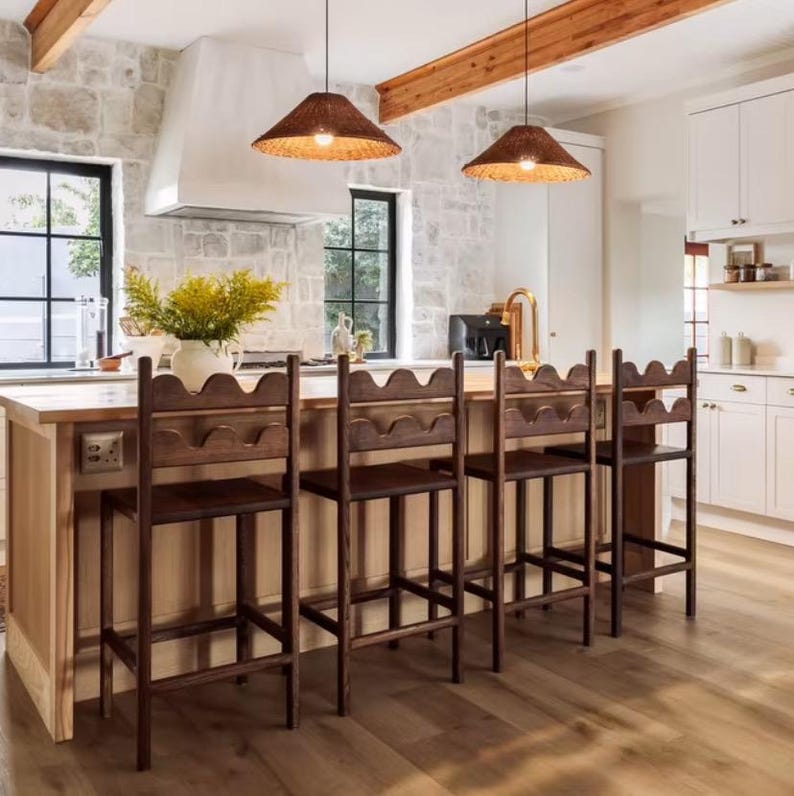 K&ouml;nnte beinhalten: Vier dunkelbraune Holz-Barhocker mit gewellten R&uuml;ckenlehnen, die an einer K&uuml;cheninsel aus hellem Holz stehen. Die Hocker haben gerade Beine und ein schlichtes Rahmendesign und befinden sich in einer hellen K&uuml;che mit Steinw&auml;nden und zwei Lampen.