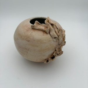 May include: A spherical ceramic vase with a smooth, off-white and light brown mottled finish. The vessel features a small circular opening at the top and a decorative, ruffled, clay-like texture applied to the right side of the exterior surface.