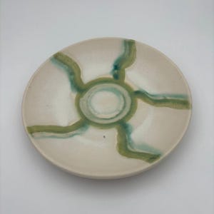 May include: A small, off-white ceramic plate with a hand-painted design. The design features a central teal circle with radiating green lines. The plate has a slightly raised edge and a smooth surface. The overall aesthetic is minimalist and artistic.
