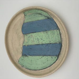 May include: A handmade ceramic plate with a beige rim and a central design of blue and green horizontal stripes. The plate has a unique, organic shape and a textured surface, perfect for serving food.
