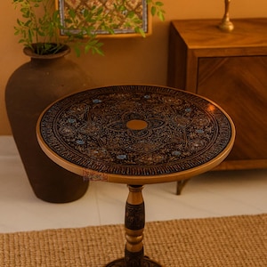 Könnte beinhalten: Ein runder, aufwendig geschnitzter Beistelltisch aus Holz mit einer dunkelbraunen Tischplatte und einem hellbraunen Untergestell. Die Tischplatte weist ein detailliertes Blumenmuster mit türkisfarbenen Akzenten auf. Der Tisch ist ca. 61 cm hoch.