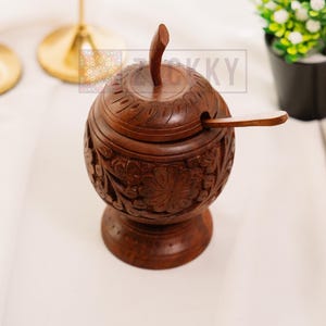 May include: A dark brown, hand-carved wooden sugar bowl with a lid and spoon. The bowl features intricate floral carvings and sits on a pedestal base. The background includes gold candle holders and a small potted plant.
