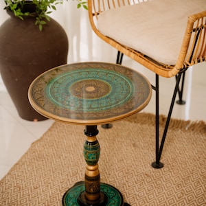 Puede incluir: Pequeña mesa auxiliar redonda con un detallado diseño de mandala turquesa y dorado en la parte superior. La mesa tiene una base de pedestal negra y dorada y está sobre una alfombra estampada. Una silla de mimbre en el fondo.
