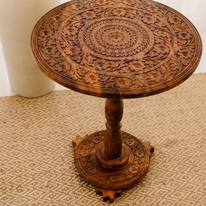 May include: A round, brown wooden side table with intricate floral carvings. The table has a pedestal base and a detailed, carved top. The table is approximately 50 cm in diameter and stands about 61 cm tall. The table is set on a woven rug.