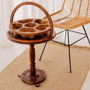 May include: A handcrafted wooden serving tray with a handle, featuring multiple compartments. The tray sits atop a decorative wooden stand with carved details. A woven chair with black metal legs is in the background. The tray is approximately 12 inches in diameter.