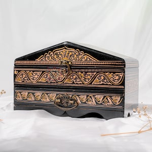 May include: Black wooden jewelry box with a decorative floral design in gold. The box has a hinged lid and a drawer, secured with a metal clasp and handle. The box is on a white surface.