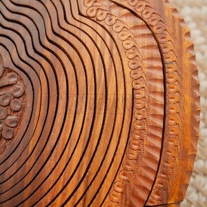May include: Close-up of a wooden fruit basket with a carved design. The basket is made of light brown wood and features a series of curved, grooved lines. The edges are decorated with a repeating pattern of small circles and lines.